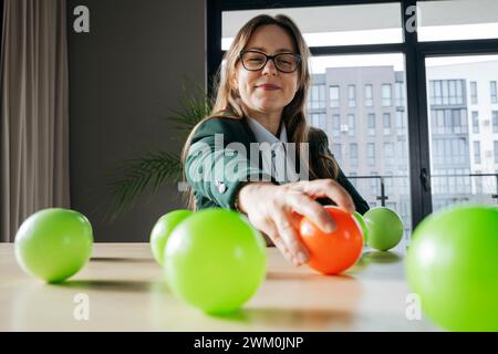 Smiling businesswoman reaching for red ball kept on desk in office Stock Photo