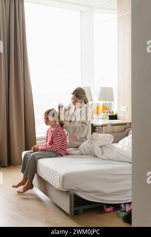 Mother tying daughter's hair sitting on bed at home Stock Photo