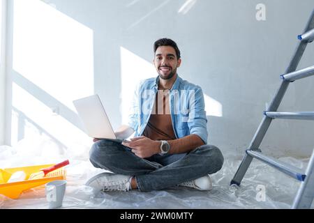 Smiling man sitting on floor with laptop in room under renovation Stock Photo