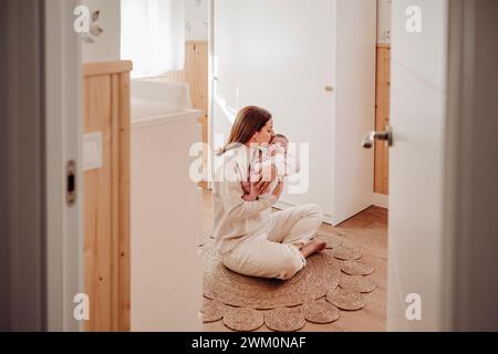 Mother sitting on wicker rug and kissing baby girl at home Stock Photo