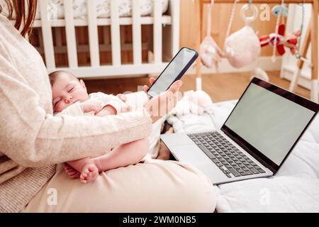 Freelancer using smart phone with baby girl sleeping near laptop at home Stock Photo