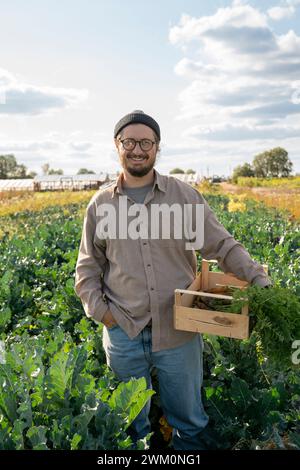 Smiling farmer with hand in pocket holding crate of leafy greens in farm Stock Photo