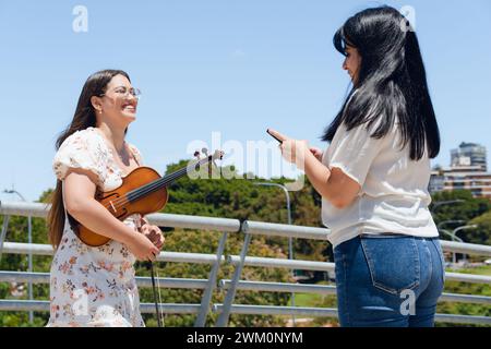 young Venezuelan Latin woman busker violinist giving her phone number to tourist fan who was listening to her play her music outdoors in Buenos Aires. Stock Photo
