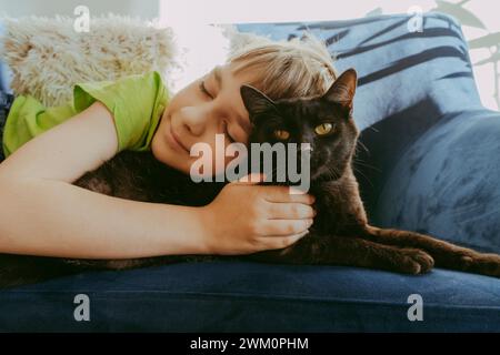 Smiling boy with eyes closed lying down with cat on sofa at home Stock Photo