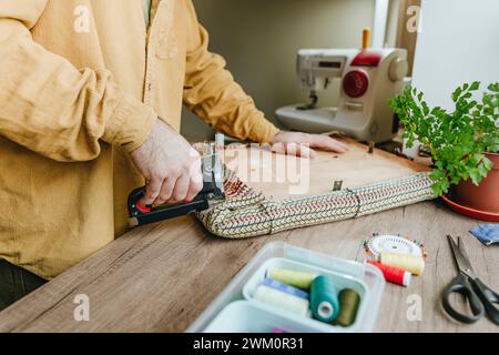 Man securing fabric from old clothes on chair with stapler at table Stock Photo