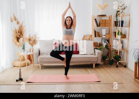 Pregnant woman practicing tree pose yoga on mat in living room Stock Photo
