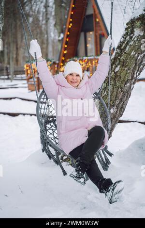 Mature woman sitting on swing near log cabin in winter Stock Photo