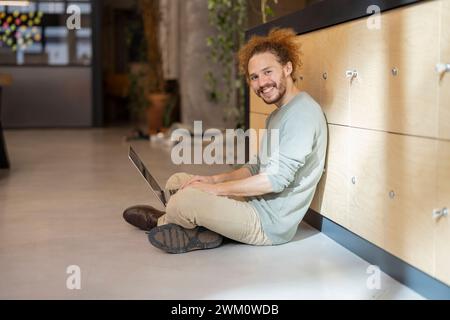 Smiling businessman using laptop sitting cross-legged on floor at workplace Stock Photo
