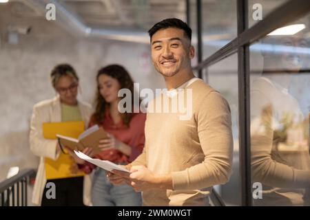 Smiling young businessman standing with document near colleagues discussing in illuminated corridor Stock Photo