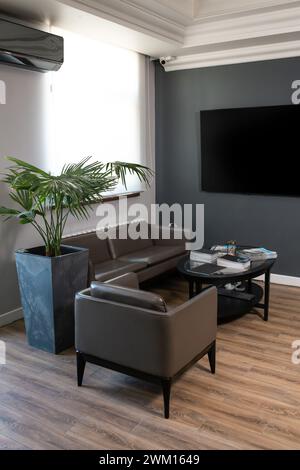 A stylish and modern office interior with dark gray walls, a large window and a monitor on the wall. comfortable leather furniture. Stock Photo