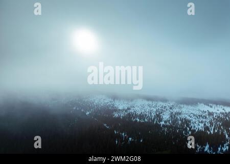 Aerial view of snowy mountain range in cloud with sun shining through during frosty day. Moody winter scenery. Stock Photo