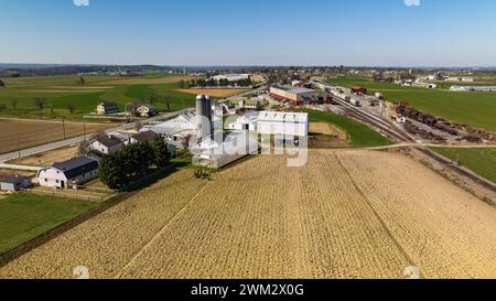 Aerial View Of A Farm Complex With Multiple Barns, Silos, And Plowed Fields In A Rural Setting Under A Clear Blue Sky. Stock Photo