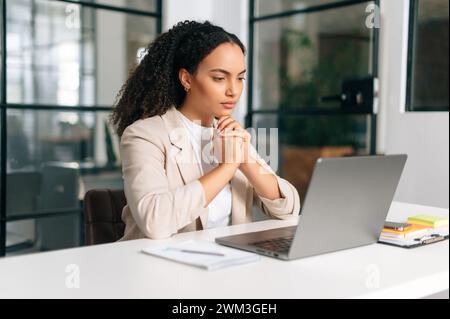 Serious busy brazilian or hispanic curly haired business woman, company ceo, hr manager, sitting at a work desk in a modern creative office, working in a laptop, focused studying information Stock Photo