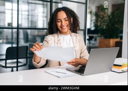 Office routine. Positive pretty brazilian or hispanic business woman, company employee, financial ceo, sitting at a work desk in modern office, working in a laptop, studying financial documents, smile Stock Photo