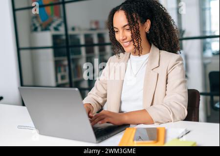Positive gorgeous successful business lady working on laptop in office, brazilian businesswoman in formal jacket sitting at a work desk in a modern space, working on a project in a laptop, smiling Stock Photo