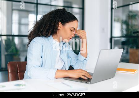 Exhausted overworked brazilian or hispanic curly haired businesswoman, company employee, works on a laptop in modern office, has a headache, migraine, massages bridge nose with eyes closed, needs rest Stock Photo