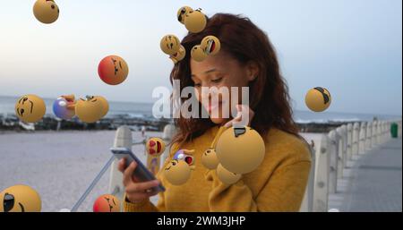 Image of emoji icons flying up with a young biracial woman smiling and using a smartphone in the b Stock Photo