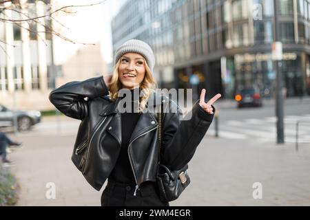 Urban positive fashion pretty girl in fashionable clothes with a stylish bag walks in the city, shows tongue and peace sign. Beauty woman on the stree Stock Photo