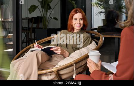 Two happy business women talking at work drinking coffee in cozy office. Stock Photo