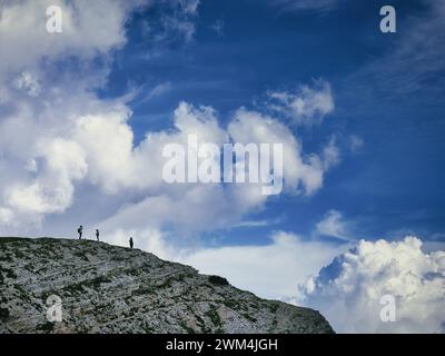 Three tourists standing on the horizon of a rocky mountain under dramatic clouds in the Dolomites, Italy, tiny human figures in the landscape. Stock Photo
