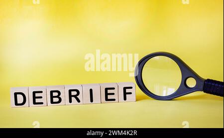 Wooden cubes with debrief text on a yellow background next to a magnifying glass Stock Photo