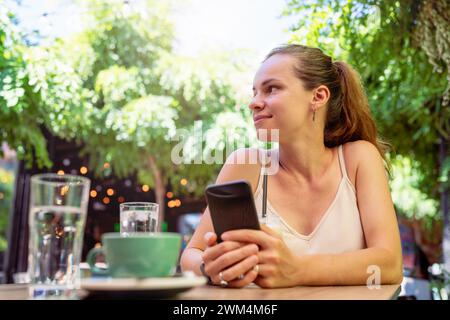 Brunette woman sits at the table at cafe holds smartphone in hands and smiling. Stock Photo