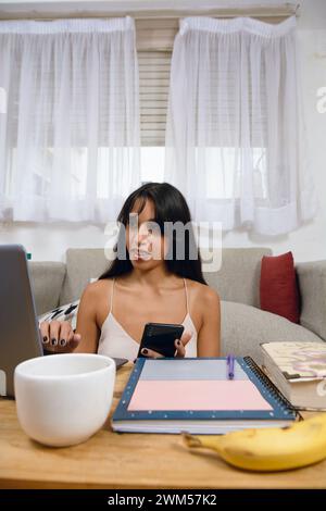 Vertical image of young Latina brunette entrepreneur at home sitting in living room using her laptop and phone, managing her business, using internet Stock Photo