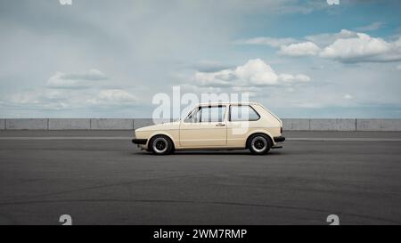 Side profile view of classic Volkswagen Golf hatchback. Beige Volkswagen Golf mk1 standing on big empty parking lot with cloudy sky in the background. Stock Photo