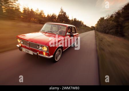 classic VAZ 2103 sedan from 1970s driving on narrow road. Three quarter front view motion shot of red Lada at sunset. Stock Photo