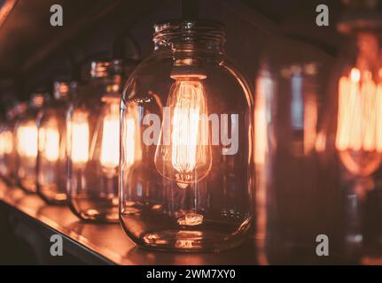 Row of light bulbs inside glass cans. DIY Edison type lamps emitting warm light on a shelf. Stock Photo