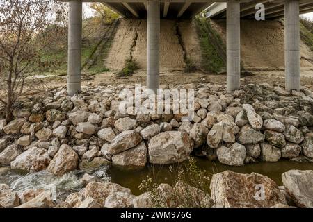 Bottom of a road bridge with a small stream passing underneath surrounded by large retaining rocks Stock Photo