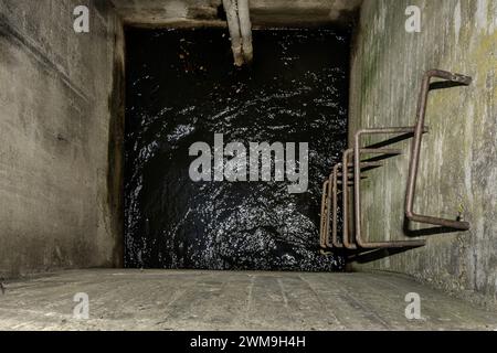 An irregular metal ladder in a square concrete structure leading down to a well filled with water Stock Photo