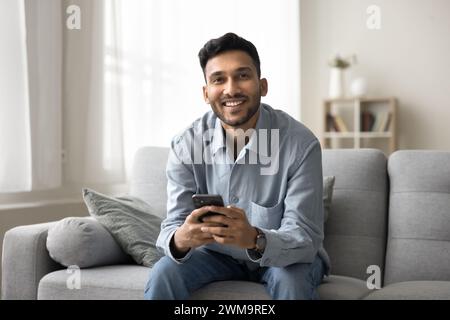 Smiling Indian man sitting on couch with smart phone Stock Photo