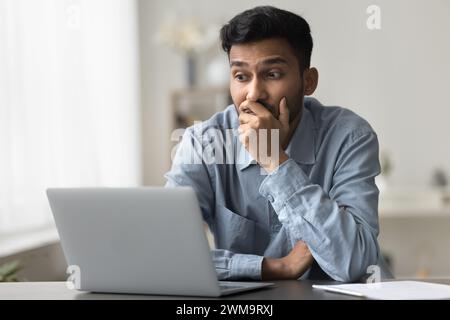 Bewildered Indian man sit at desk looking at laptop screen Stock Photo