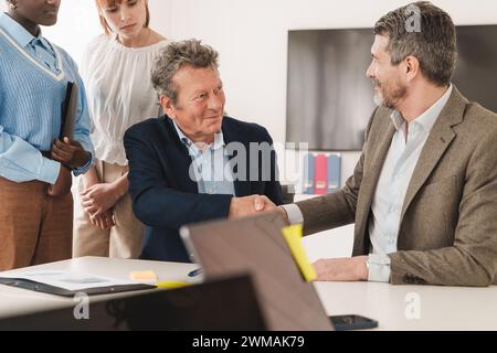 Two businessmen shaking hands in office with team watching - Agreement and partnership concept with multiethnic colleagues Stock Photo