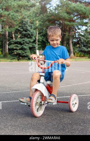 Smiling cute boy in with a crooked look is riding a red old tricycle in a park lot.z Stock Photo