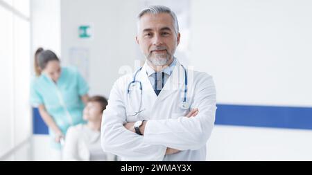 Confident doctor posing at the hospital, a nurse and a patient are talking together in the background, healthcare concept Stock Photo