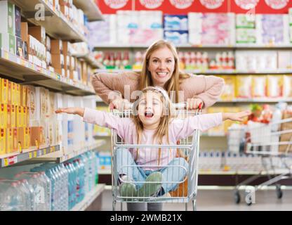 Cheerful cute girl sitting in a shopping cart at the supermarket, her mother is pushing her: grocery shopping and lifestyle concept Stock Photo