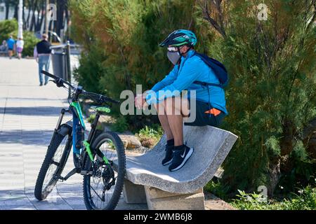 Vigo, Pontevedra, Spain; June,23,2021; MTB cyclist with glasses, helmet, mask rests sitting on a bench next to his bike Stock Photo