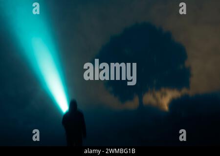 A mysterious lone hooded figure, pointing a torch beam into the sky. On a spooky foggy night in the countryside. With a grunge blurred edit Stock Photo