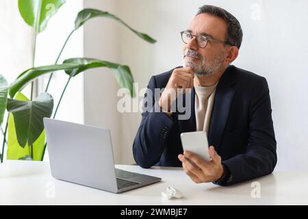 Pensive senior man with glasses holding a smartphone and looking thoughtfully into the distance Stock Photo