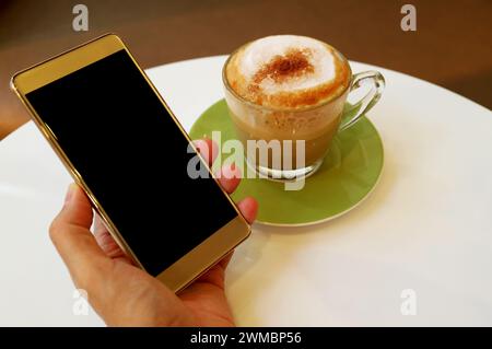 Hand Holding blank screen cellphone with a cup of cappuccino coffee in the backdrop Stock Photo