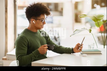 Positive stylish african american guy drinking coffee, using smartphone Stock Photo