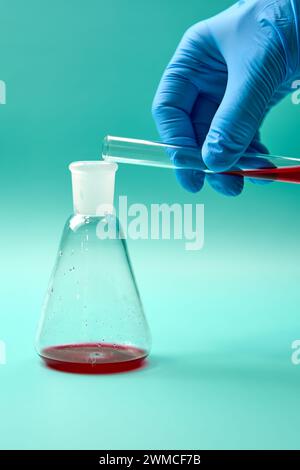 Hand of anonymous medical scientist in disposable glove pouring red liquid sample from test tube into titration flask during science laboratory resear Stock Photo