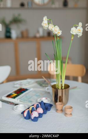 Close-up of a paintbrush, paint palette and carton of eggs being painted for Easter decorations with narcissus flowers on a table Stock Photo