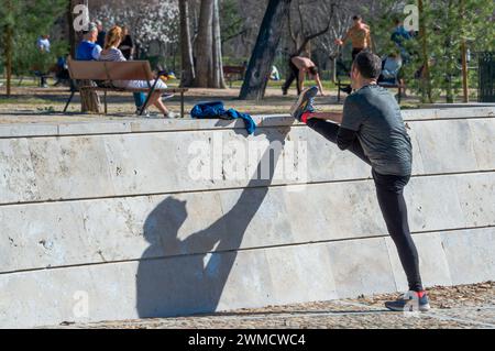 A male runner stopped to stretch his legs against a small wall in a park casting a dark shadow during a sunny day in Madrid Spain. Stock Photo