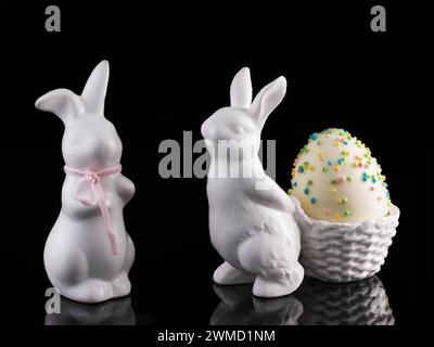 Two decorative white Poezelan Easter bunnies on a black, reflective base with a foam Easter egg Stock Photo