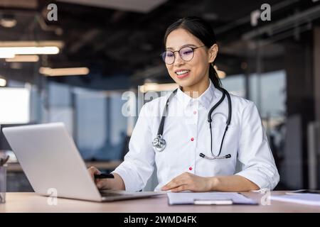 Smiling Asian female doctor in a white coat with stethoscope using laptop in a well-lit contemporary office setting. Stock Photo