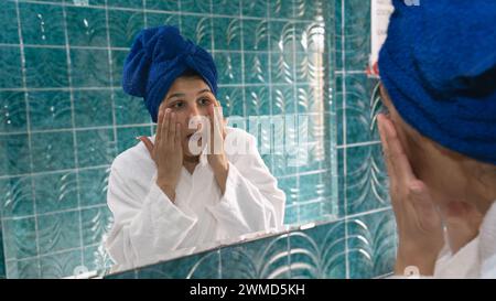 young woman in the bathroom with hands on her face in front of the mirror watching her expression lines Stock Photo