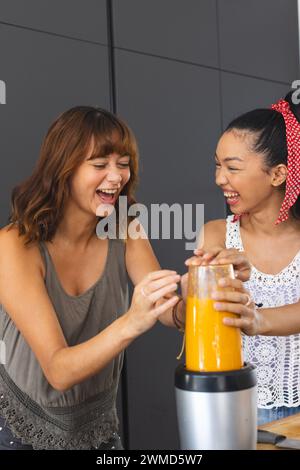Young Caucasian woman and young Biracial woman laugh together in a home kitchen, making smoothie Stock Photo